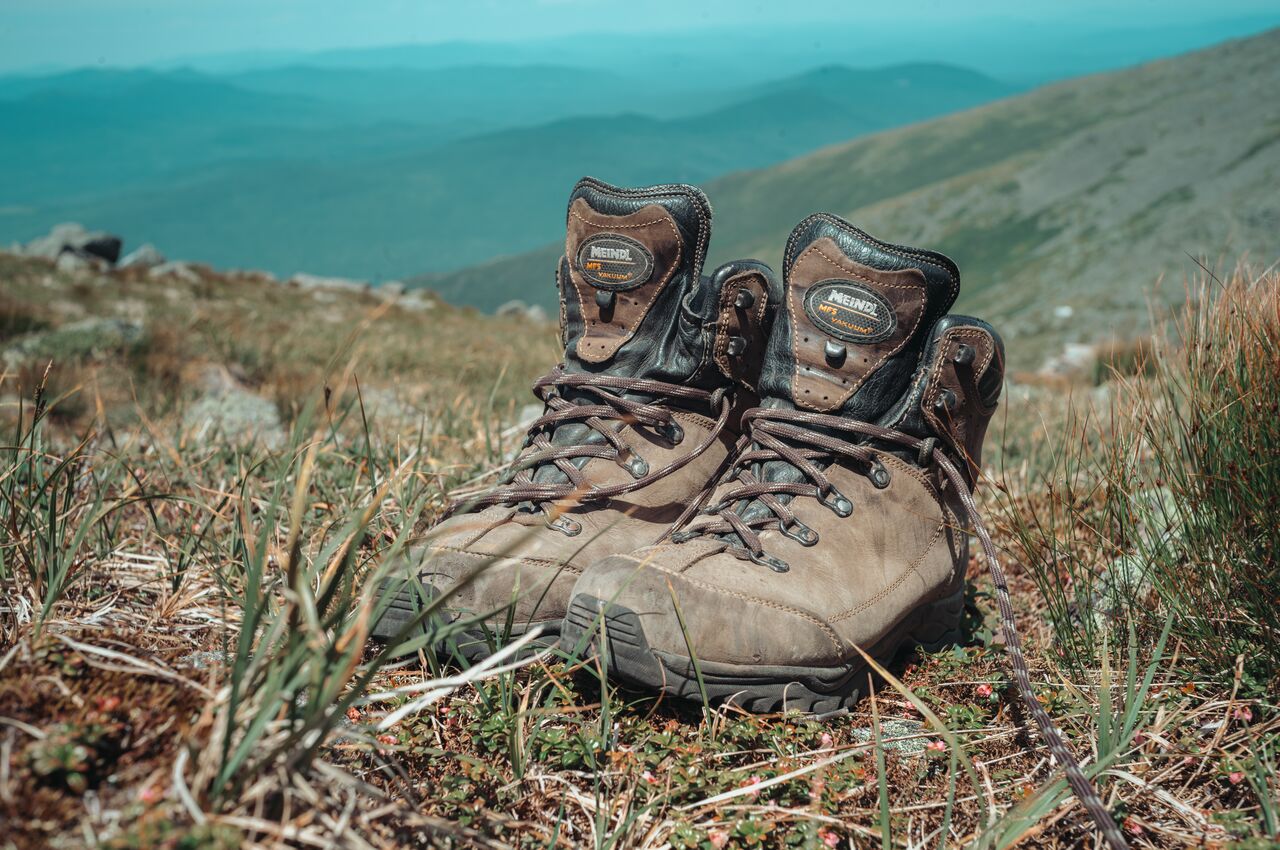 A pair of worn brown leather hiking boots rests on a grassy hillside, with mountain ranges in the distance.