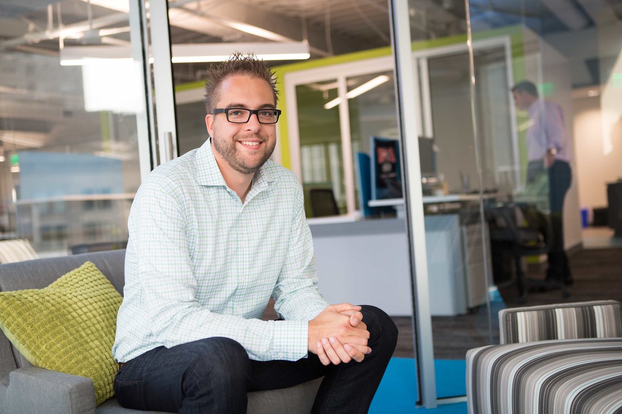 A man wearing glasses and a checkered shirt sits on a couch, smiling in a modern office setting.