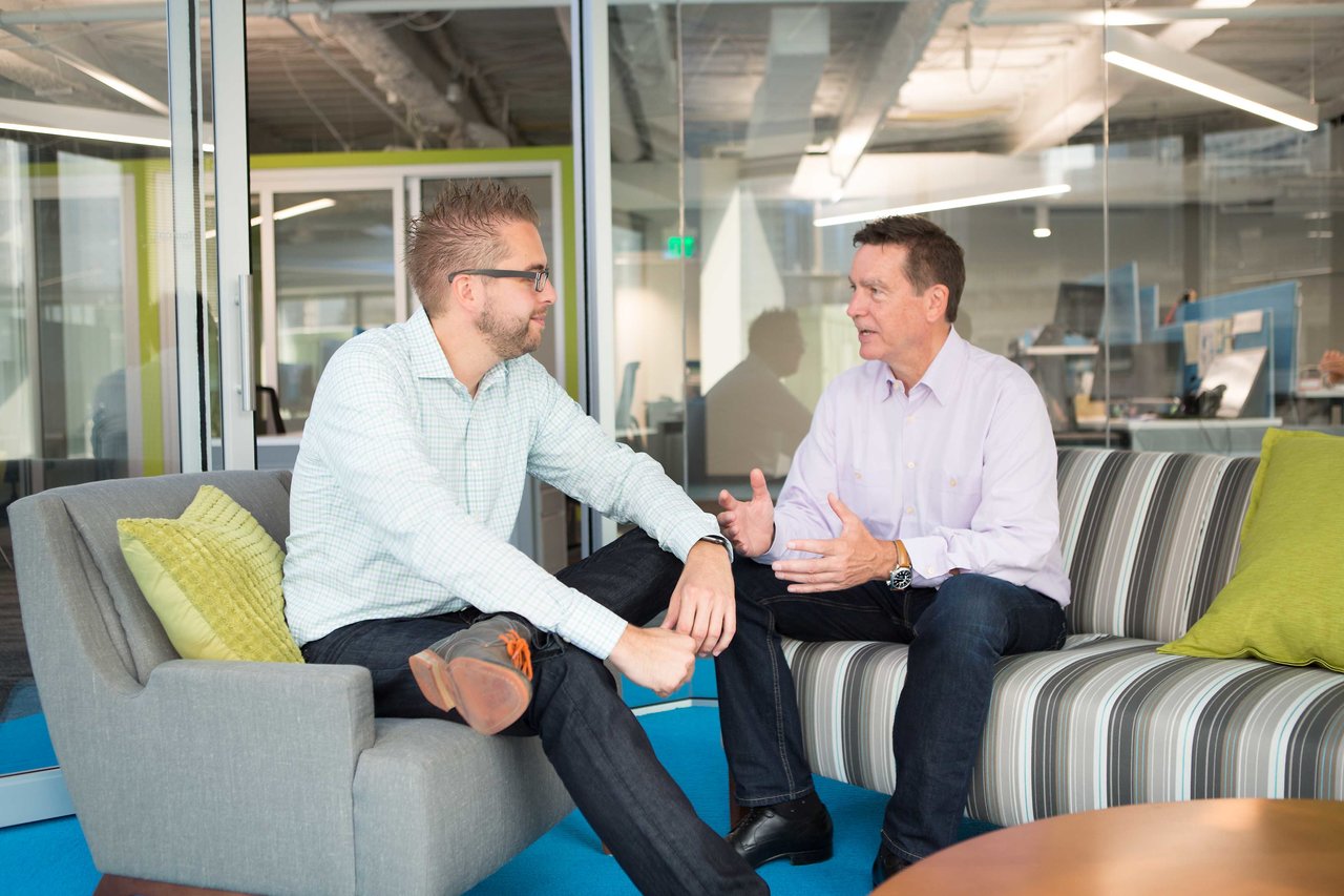 Two men sit on office lounge furniture, engaged in a conversation.