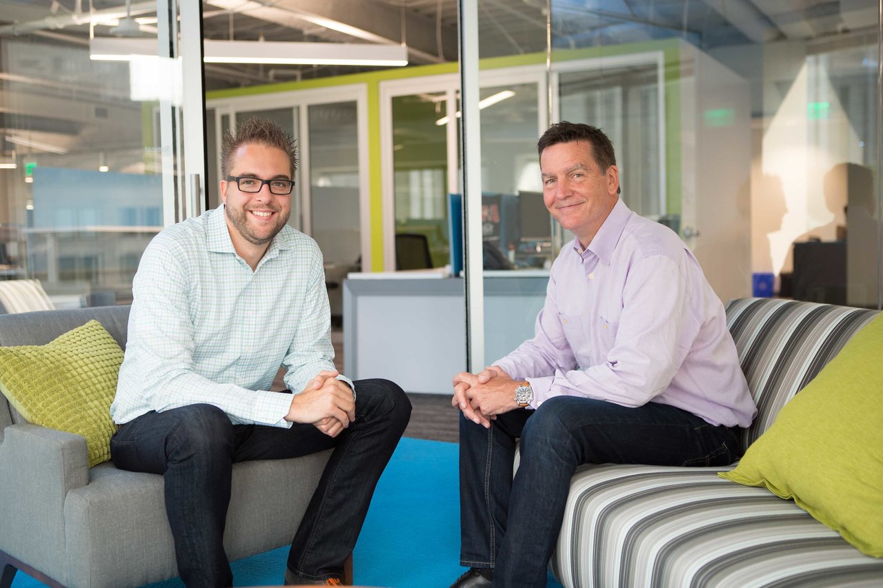 Two men sitting in a modern office lounge, smiling at the camera.