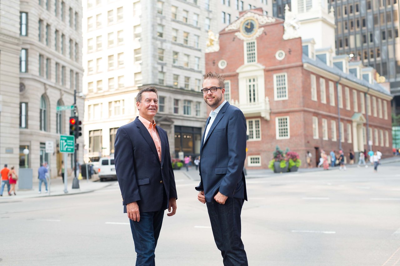 Two men in business attire stand on a city street, posing for a professional photo with historic buildings behind them.