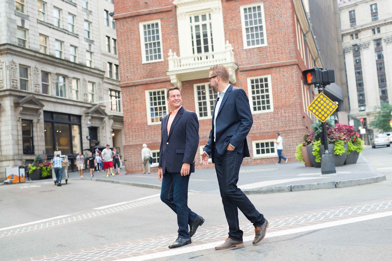 Two men in business attire walk and talk on a city street near a historic brick building.