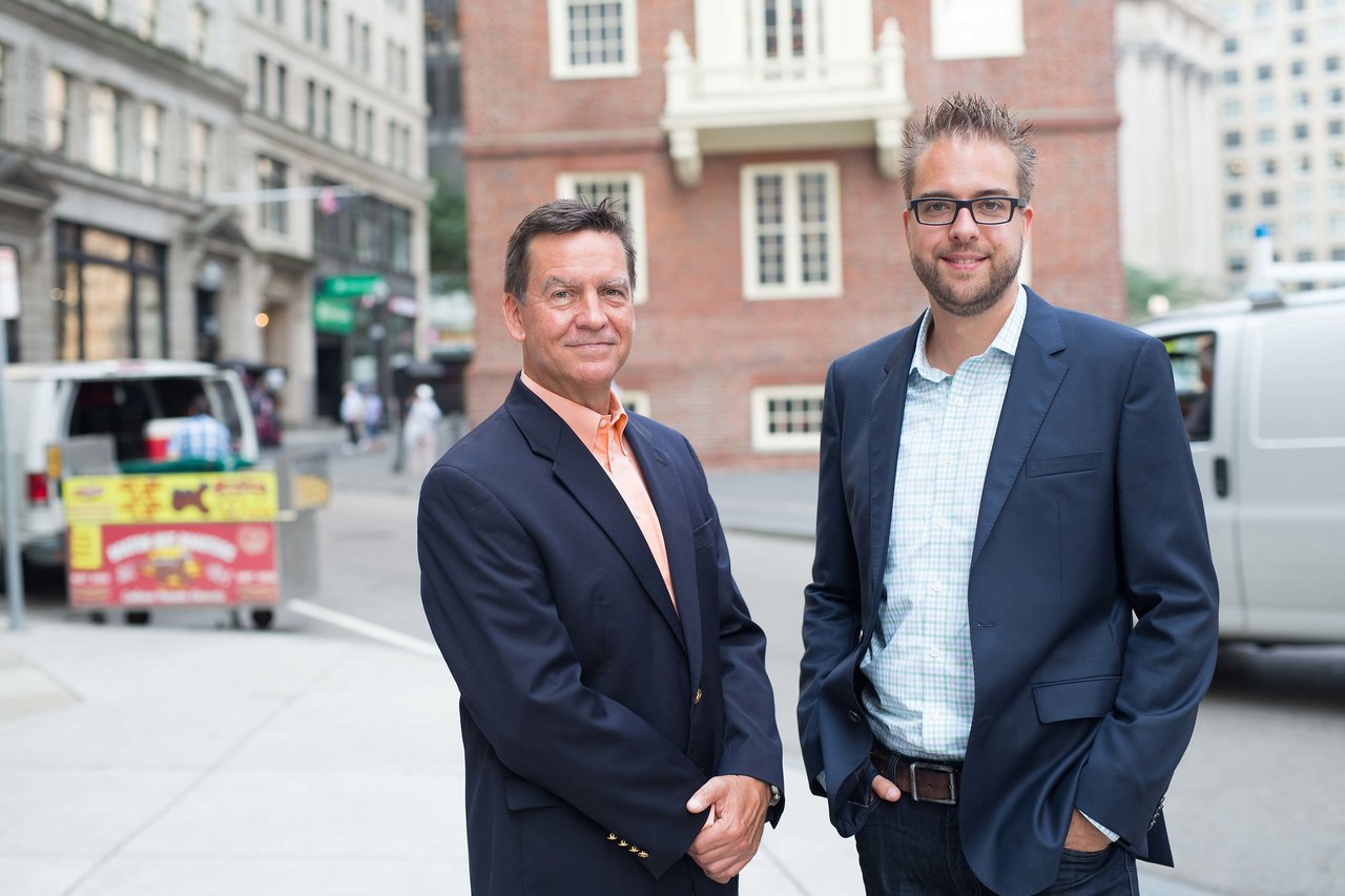 Two men in business attire stand on a city street, posing for a professional photo with a historic building behind them.
