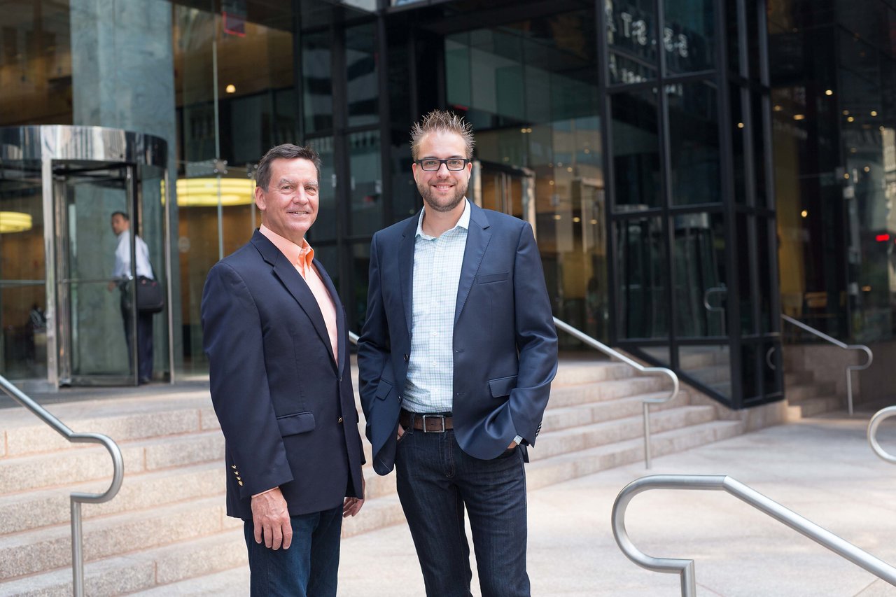 Two men in business attire stand outside a modern office building, smiling at the camera.
