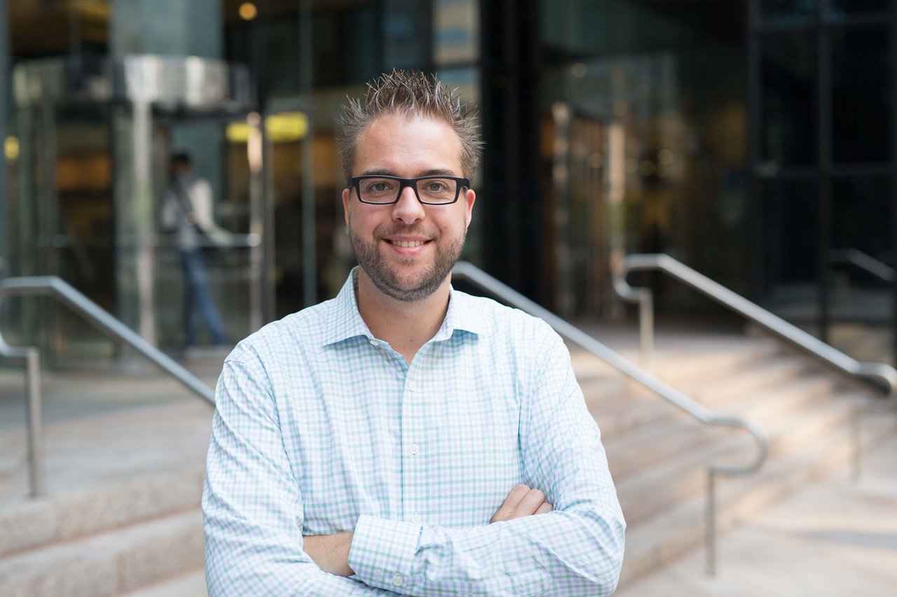 A man wearing glasses and a checkered shirt stands outside a modern building, smiling with arms crossed.