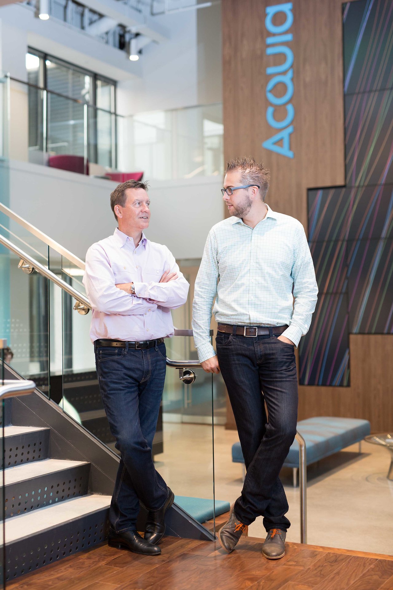Two men stand near a staircase in an office, engaged in conversation, with an "Acquia" sign in the background.