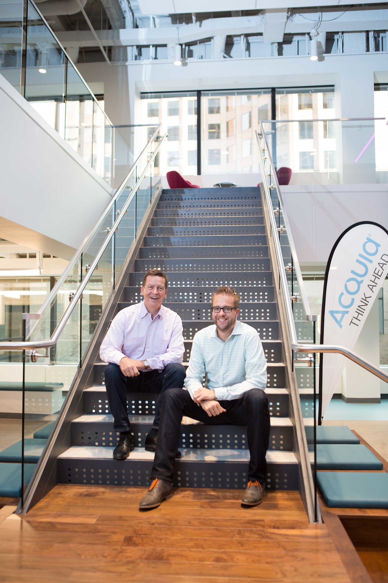 Two men sit on a modern office staircase, smiling at the camera.