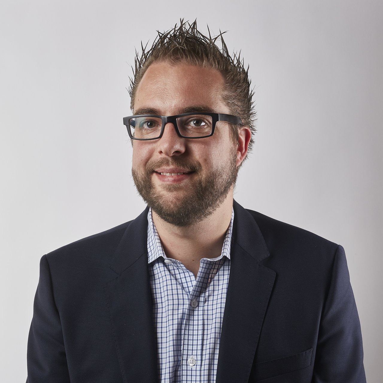 A man wearing glasses and a suit jacket smiles in a professional headshot against a plain background.