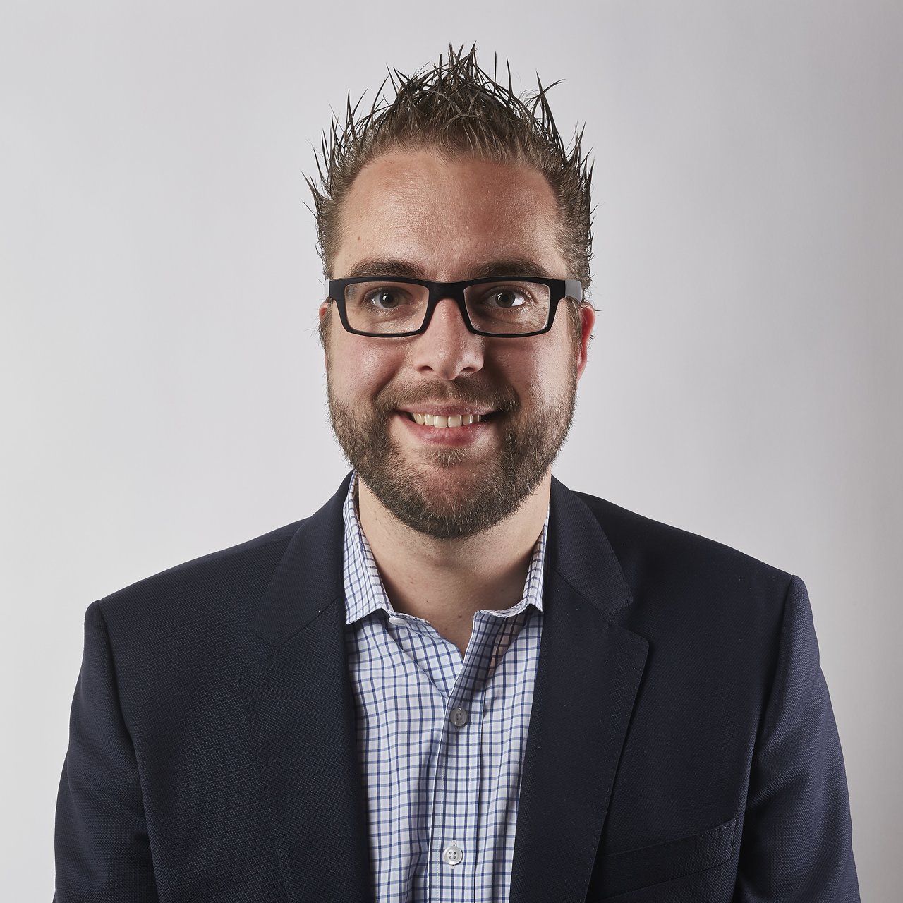 A man wearing glasses and a suit jacket smiles at the camera against a plain white background.