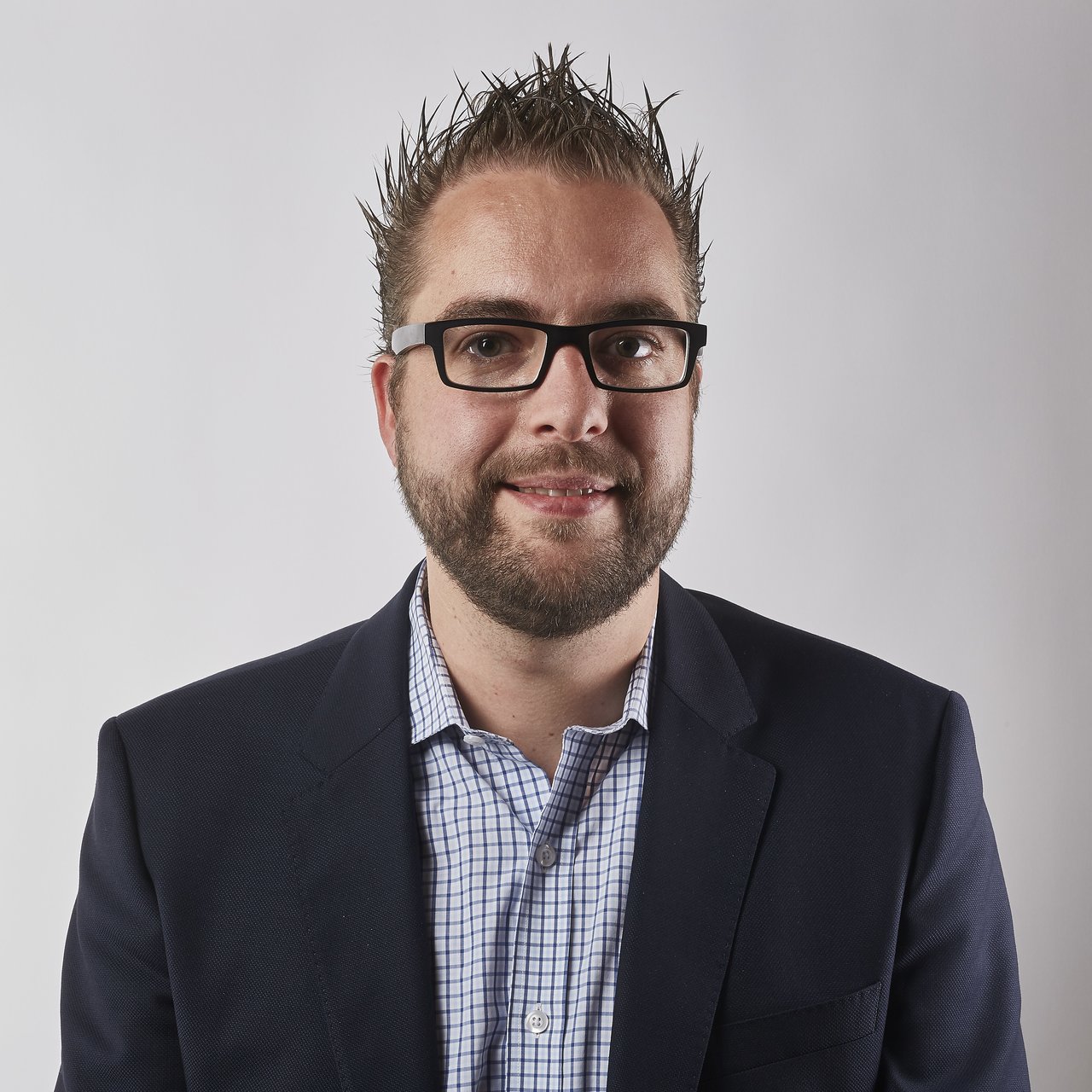 A man wearing glasses and a suit jacket smiles at the camera against a plain white background.