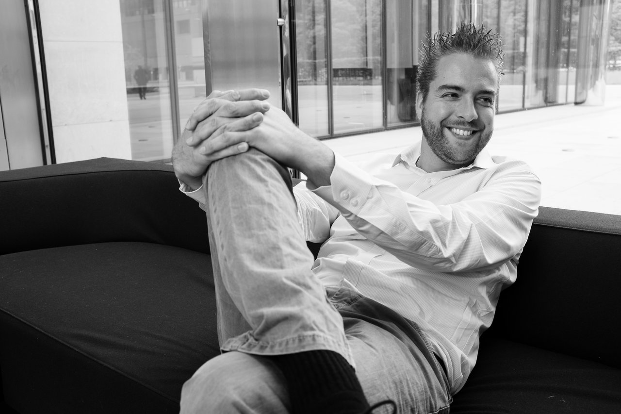 A man in a white shirt sits on a couch, smiling and relaxing with one leg crossed over the other.