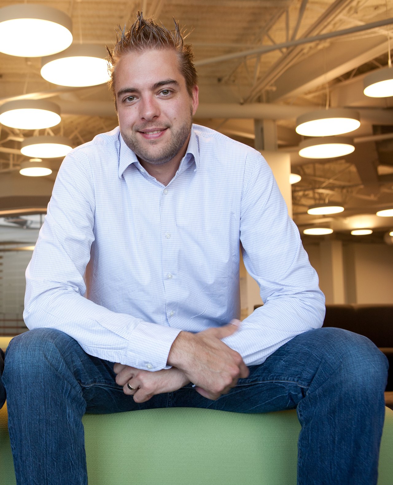 A man in a light blue shirt and jeans sits on a green bench, smiling in a modern office setting.