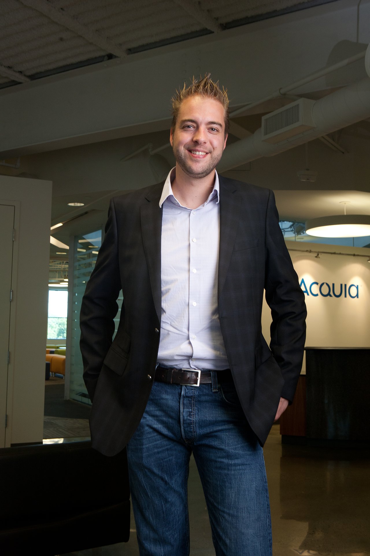 A man in a blazer and jeans stands smiling in an office with an "Acquia" sign in the background.
