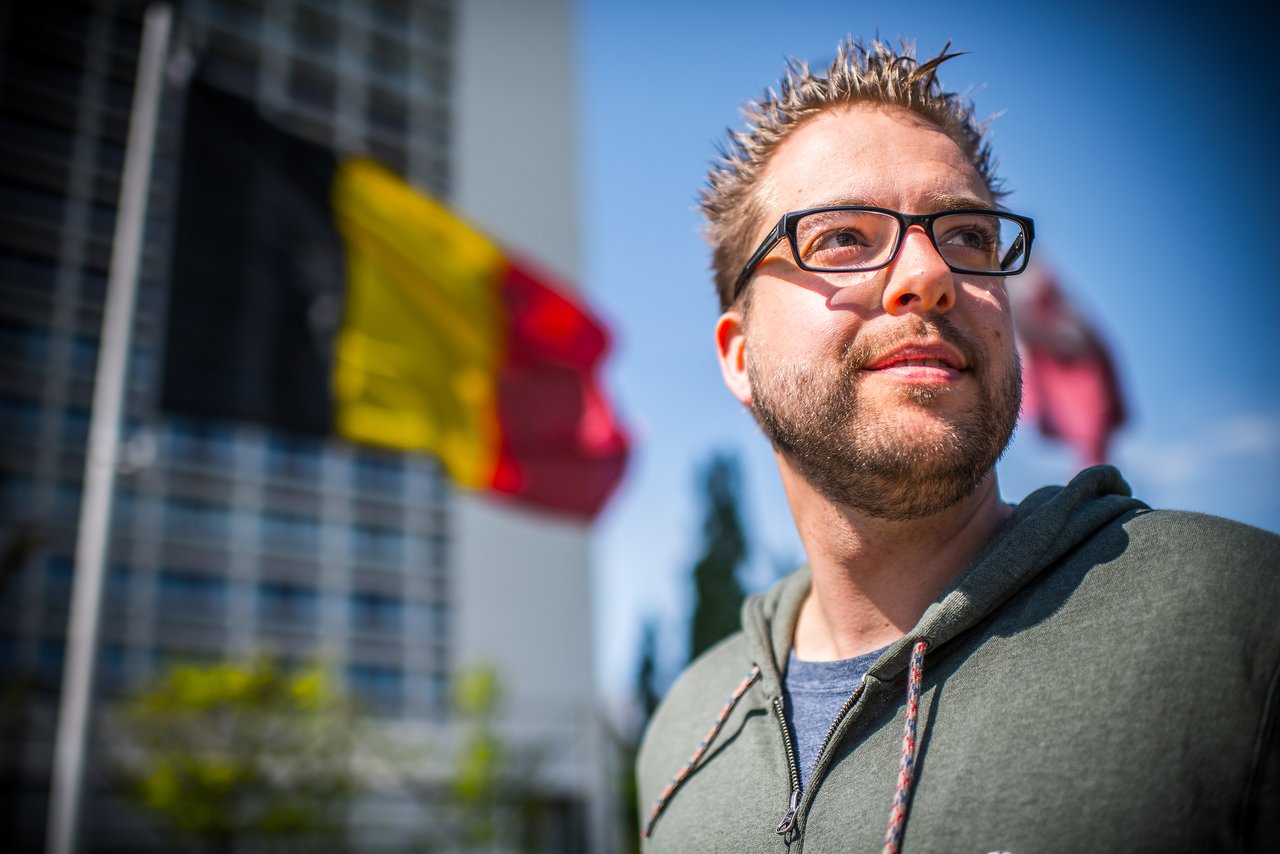 A man wearing glasses and a hoodie looks into the distance, with a Belgian flag waving behind him.