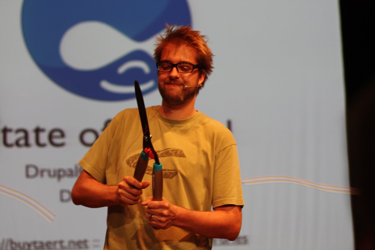 A speaker on stage at DrupalCon Paris holds garden shears while presenting in front of a large screen.