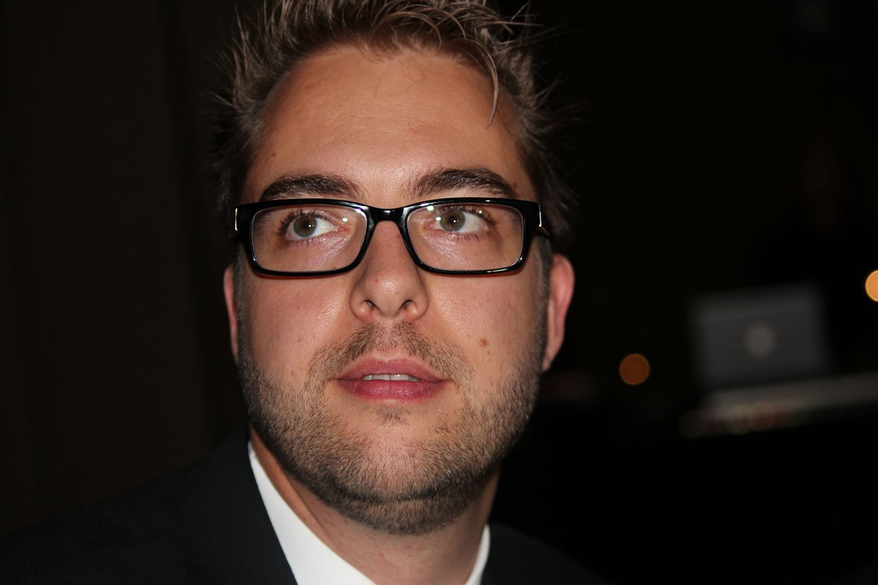 A man wearing glasses and a suit looks slightly upward at an event, with a blurred background behind him.