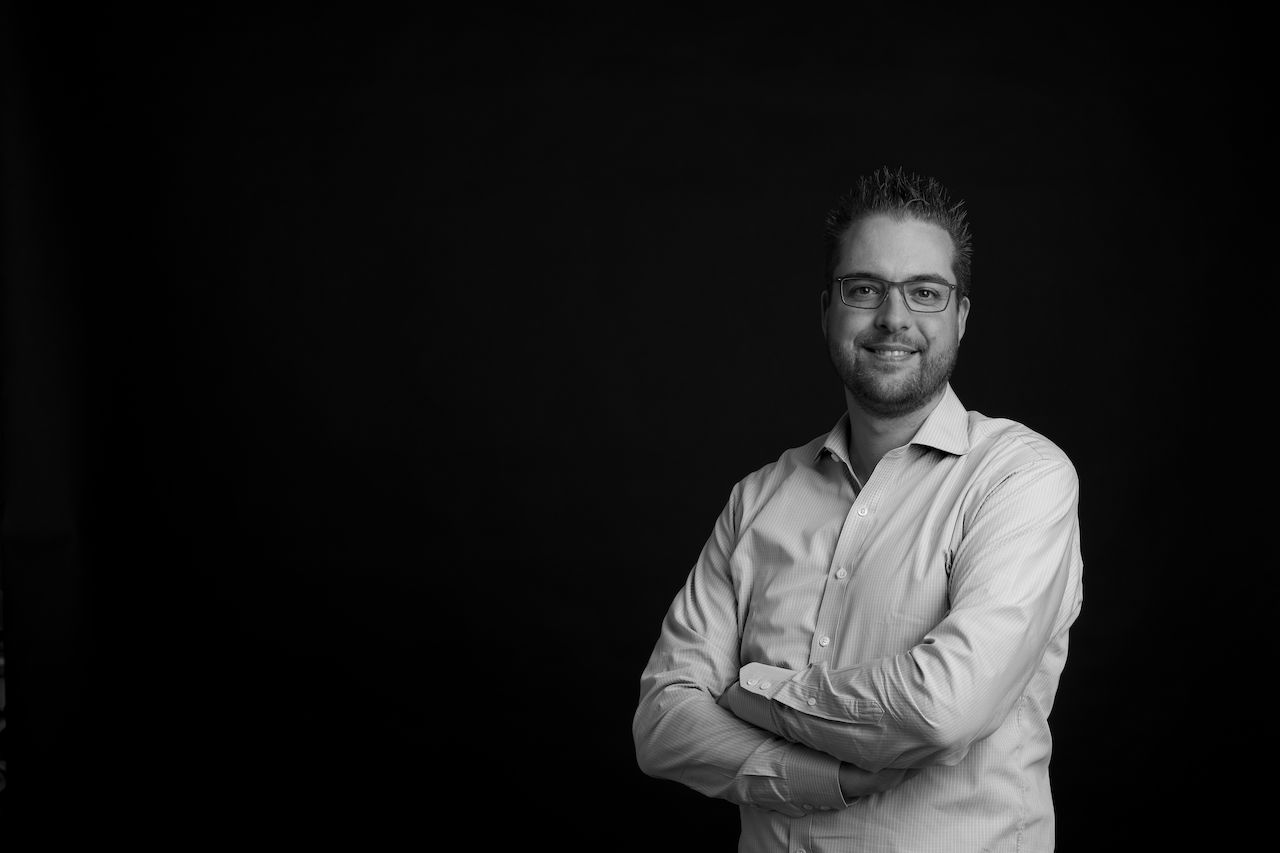 A man wearing glasses and a button-up shirt stands with arms crossed, smiling against a dark background.