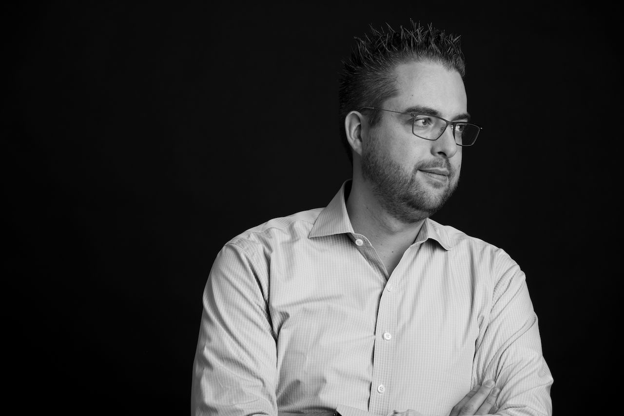 A man wearing glasses and a button-up shirt looks to the side with his arms crossed.