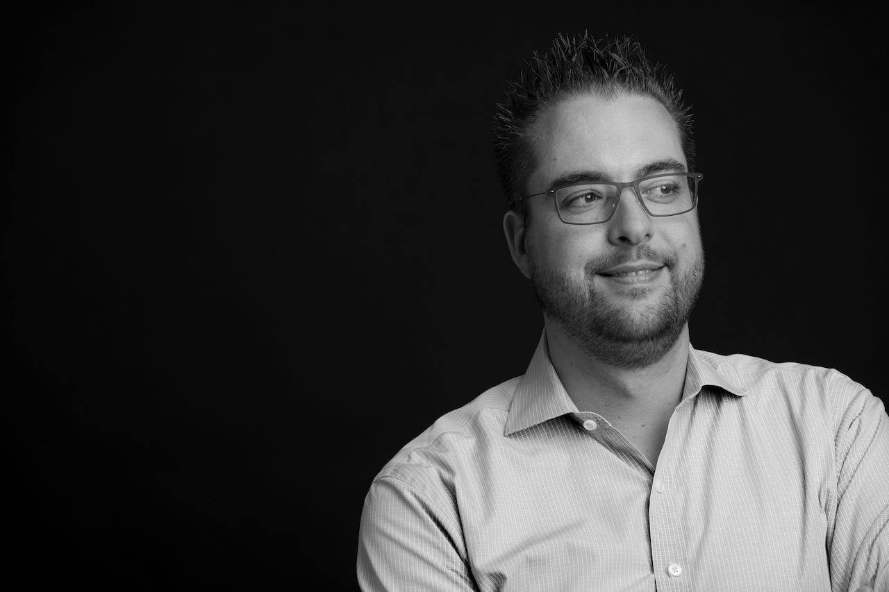 A man wearing glasses and a collared shirt looks off to the side with a slight smile.