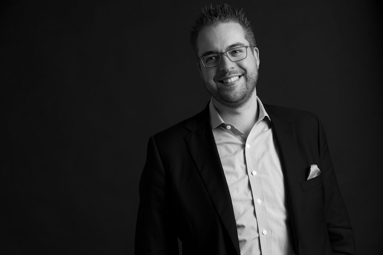 A man wearing glasses and a suit smiles at the camera against a dark background.