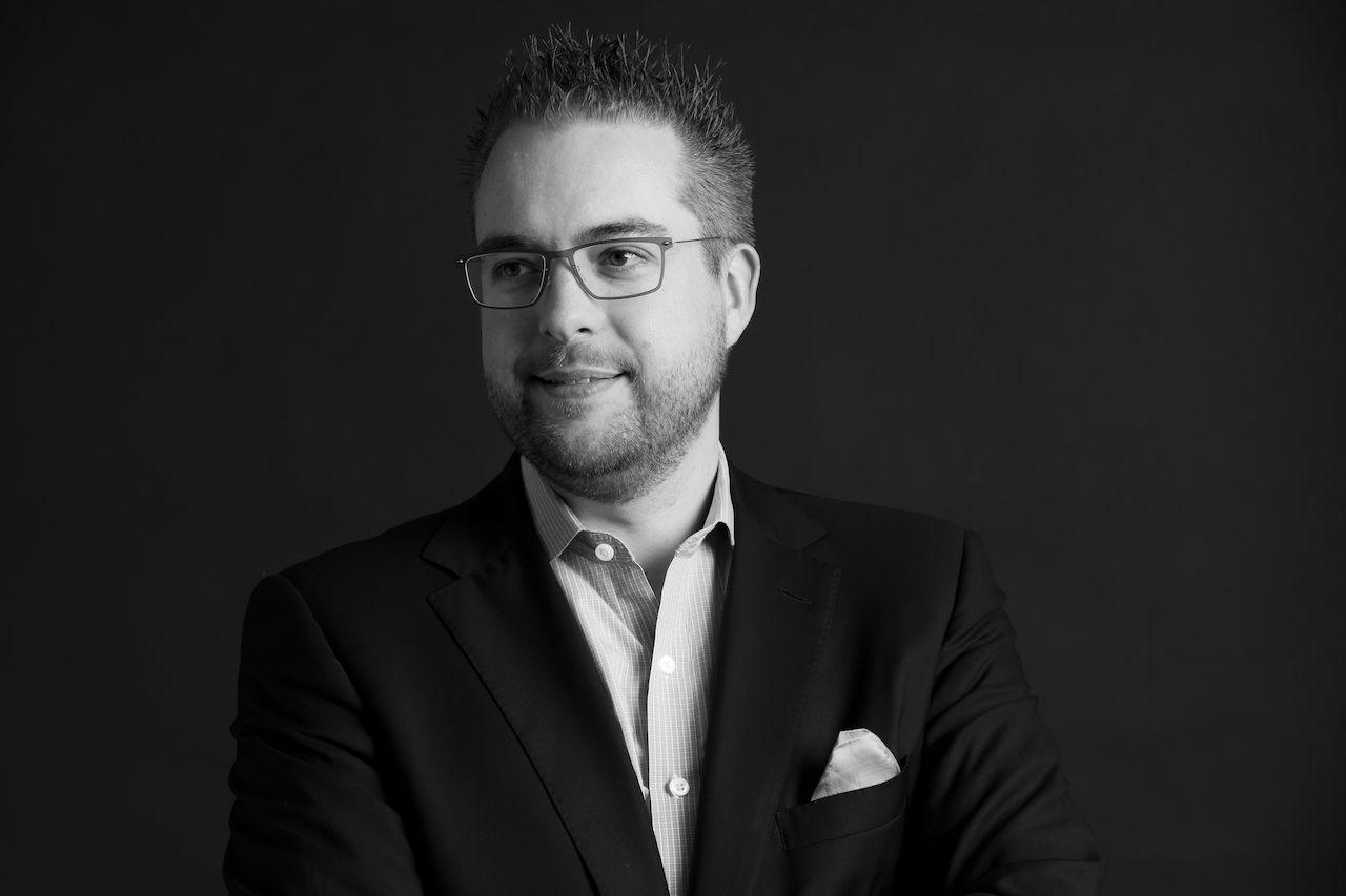A man wearing glasses and a suit looks slightly to the side with a neutral expression against a dark background.