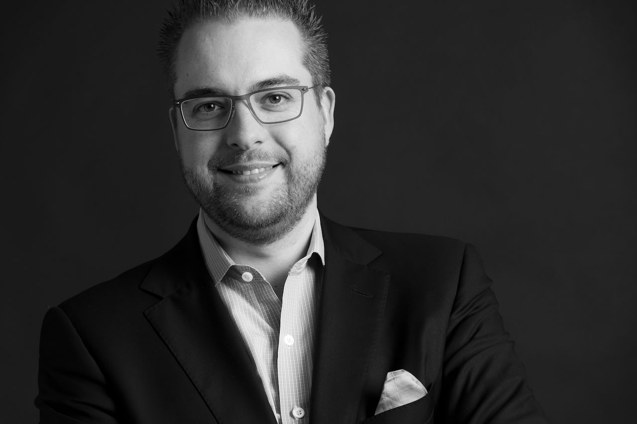A man wearing glasses and a suit smiles at the camera against a dark background.