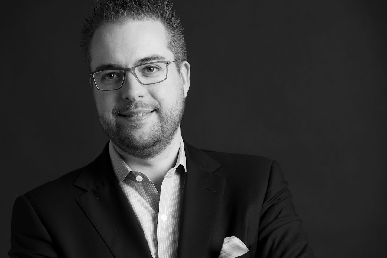 A man wearing glasses and a suit smiles slightly while posing for a professional black-and-white portrait.