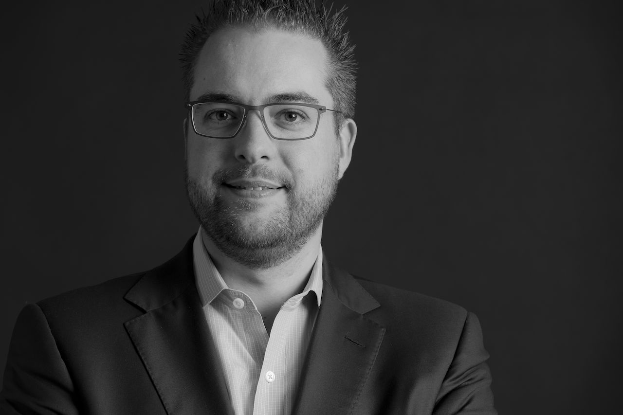 A man wearing glasses and a suit looks directly at the camera with a slight smile.
