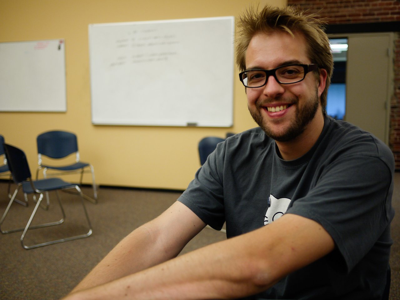 A smiling man wearing glasses and a dark t-shirt sits in a classroom with whiteboards and chairs in the background.