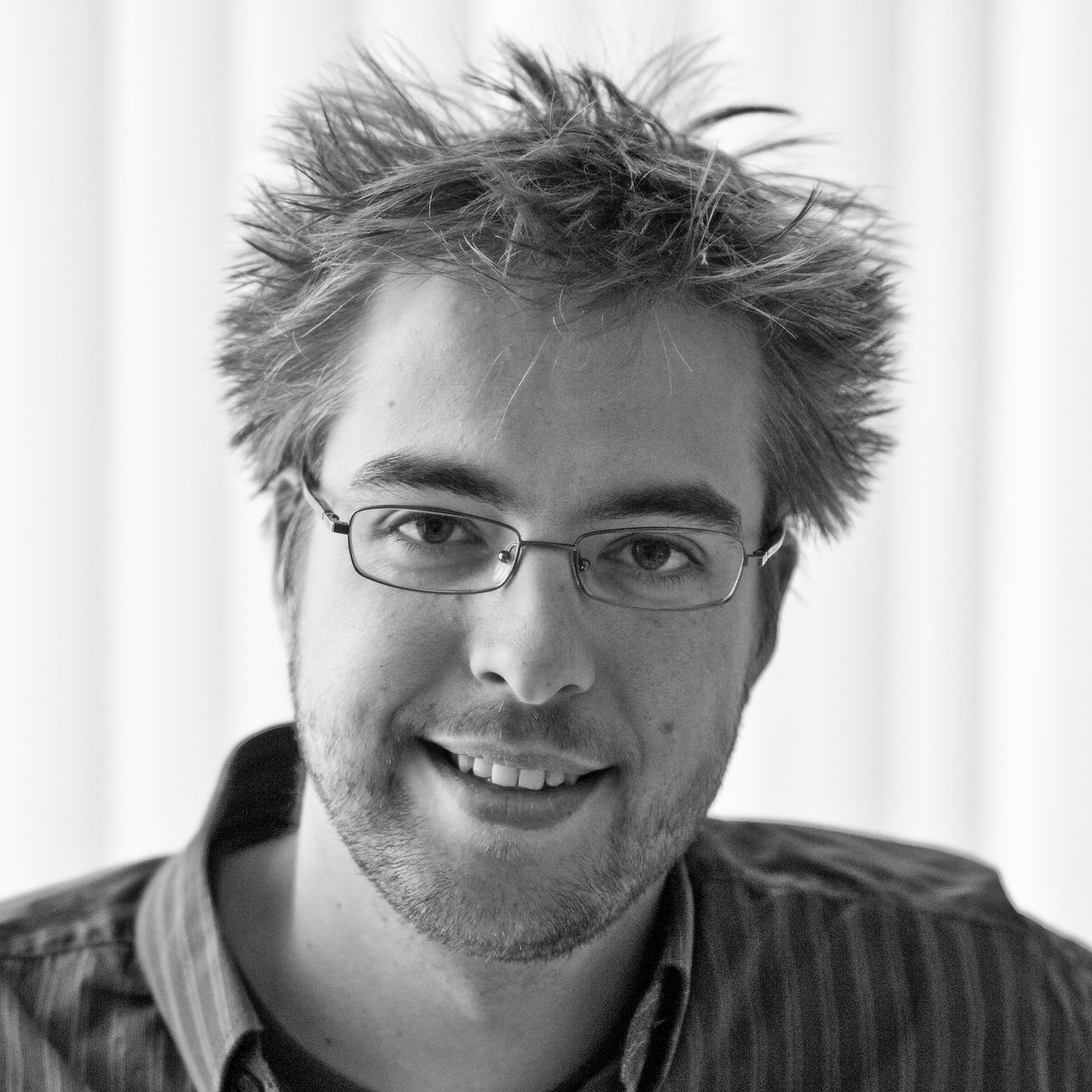 Black and white portrait of a smiling person with glasses and messy hair, wearing a collared shirt.