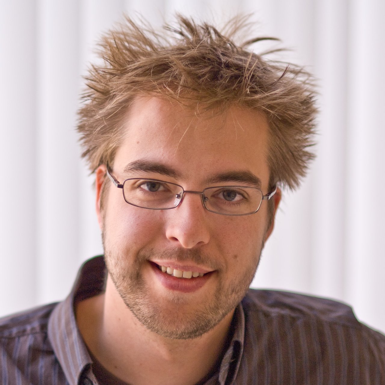 A man with glasses and messy hair smiles at the camera, wearing a dark shirt against a light background.