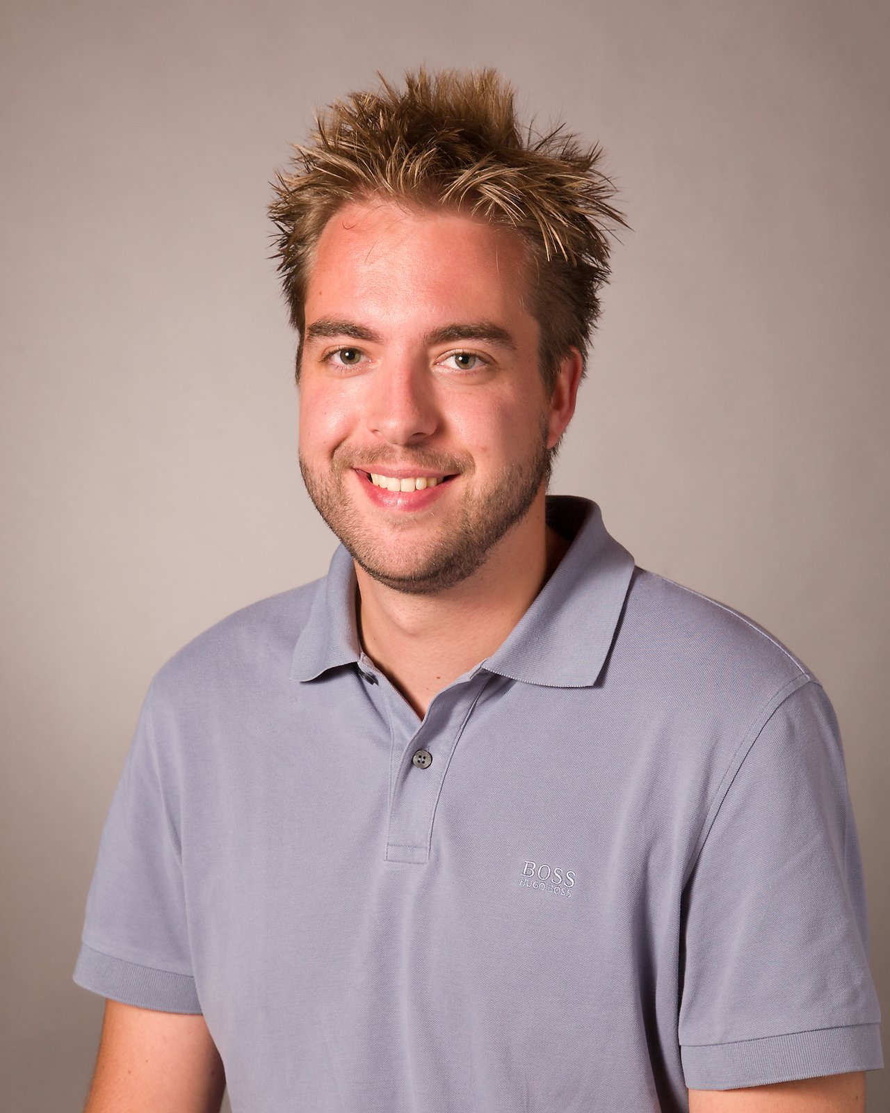 A man with short, spiky hair and a beard smiles while wearing a light gray polo shirt.