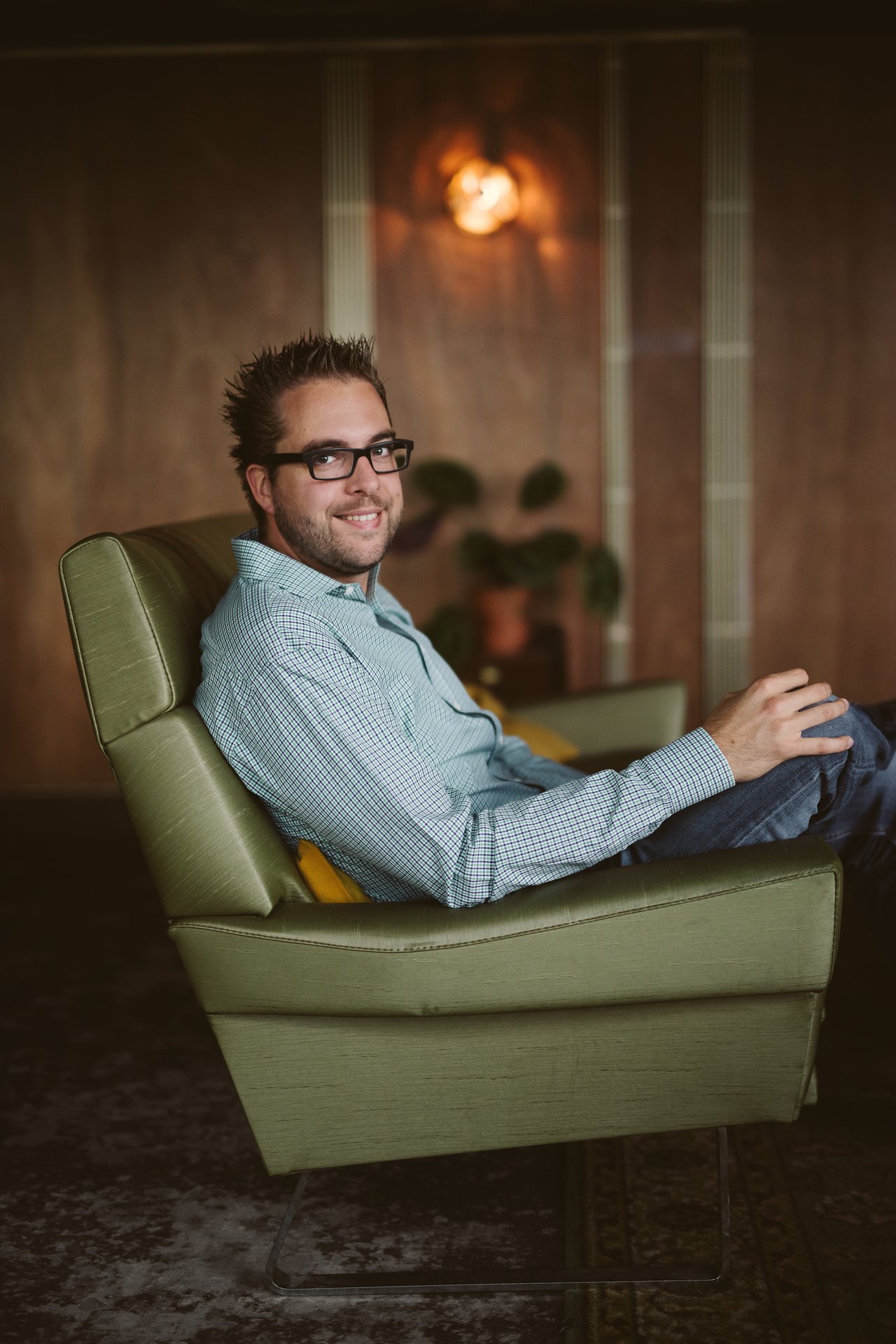 A man wearing glasses and a checkered shirt sits in a green chair, smiling at the camera.
