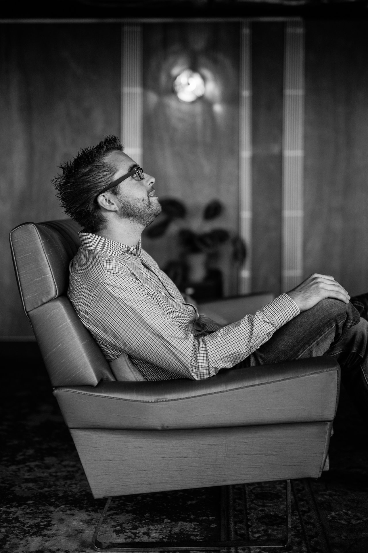 A man wearing glasses and a checkered shirt sits in a reclined chair, looking up thoughtfully.