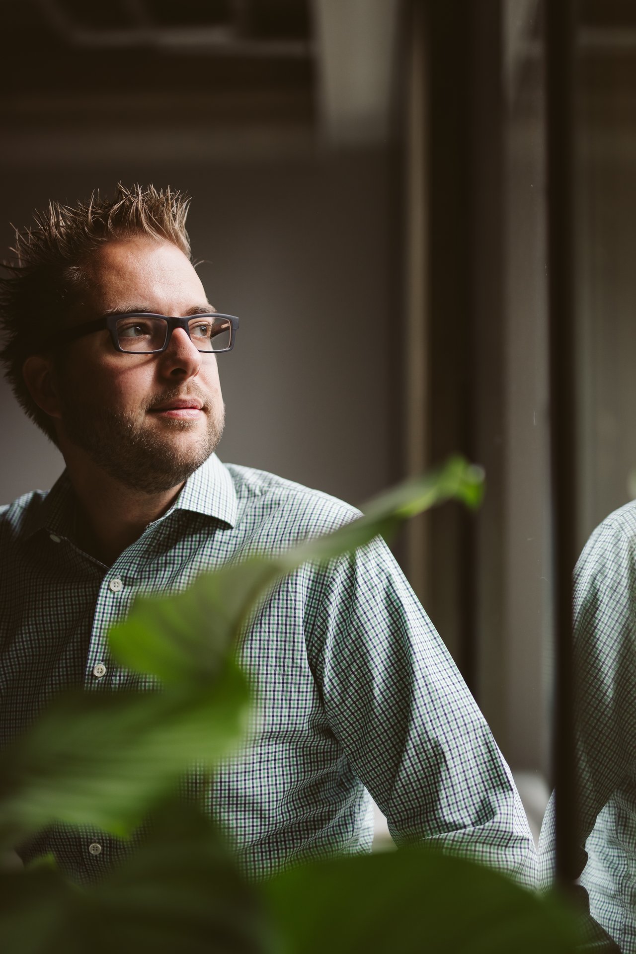 A man wearing glasses and a checkered shirt looks out the window, partially obscured by green plant leaves.
