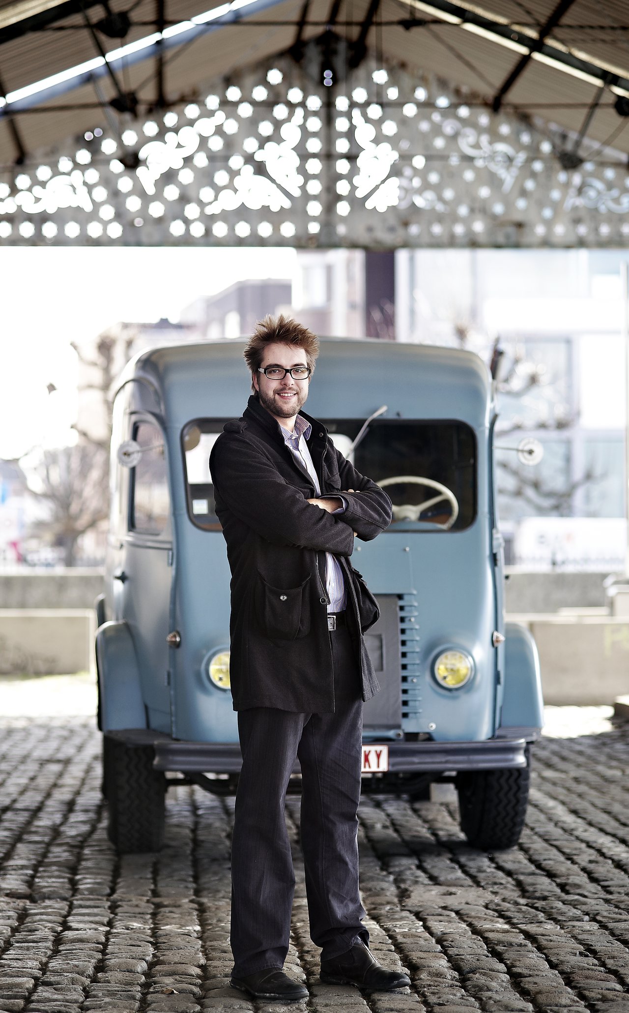 A man with glasses and a beard stands confidently with crossed arms in front of a vintage blue van.