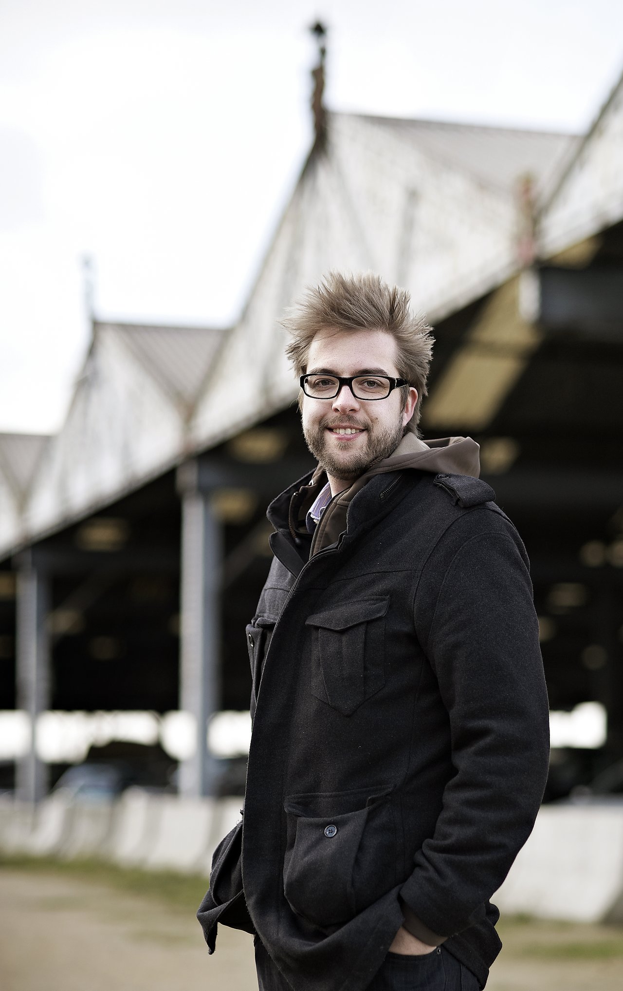 A man with glasses and a black coat stands outdoors, smiling at the camera.