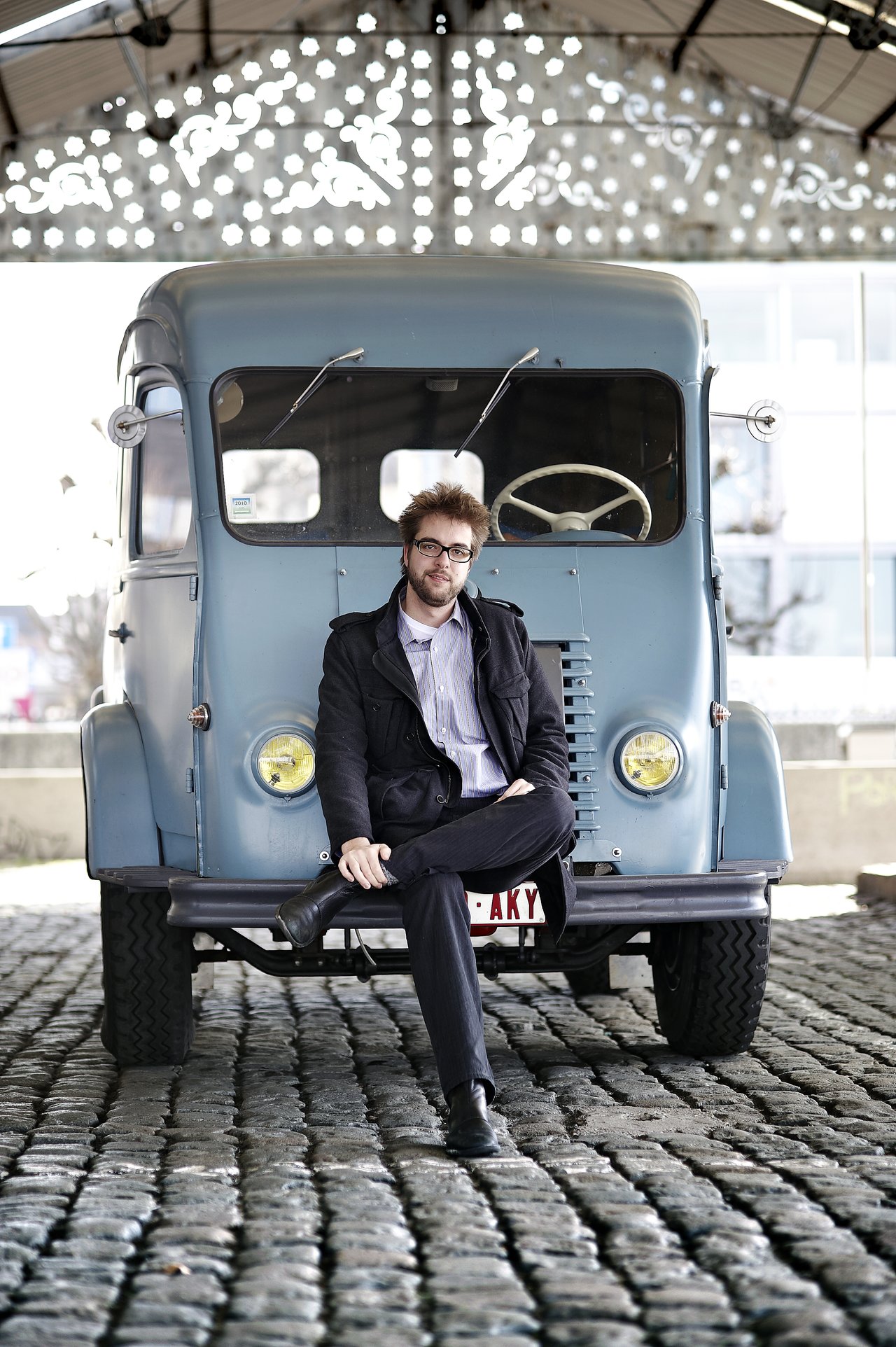A man in a suit and glasses sits casually on the front of a vintage blue truck, looking forward.