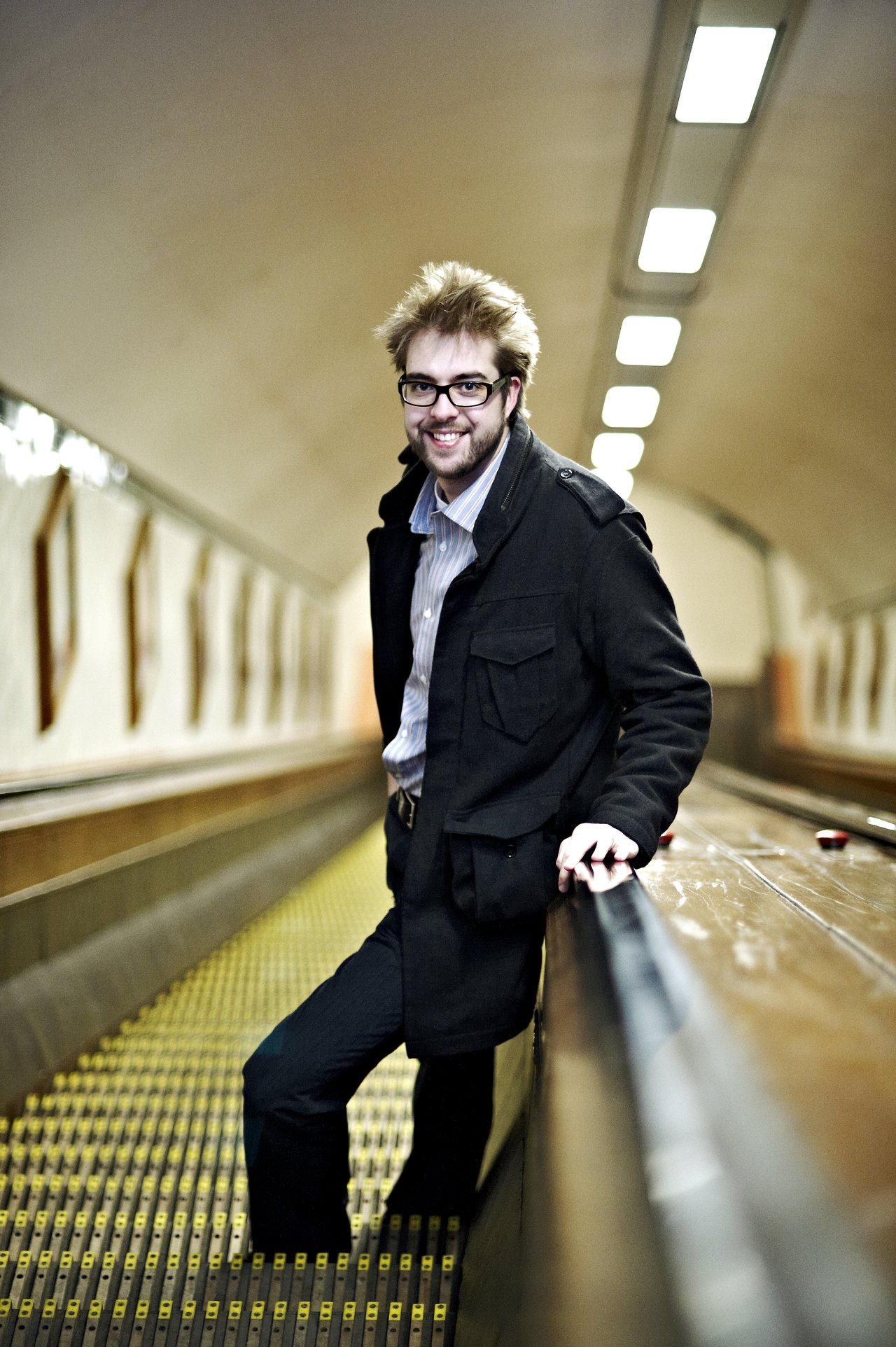 A man with glasses and a black coat smiles while leaning on a handrail in a tunnel with escalators.