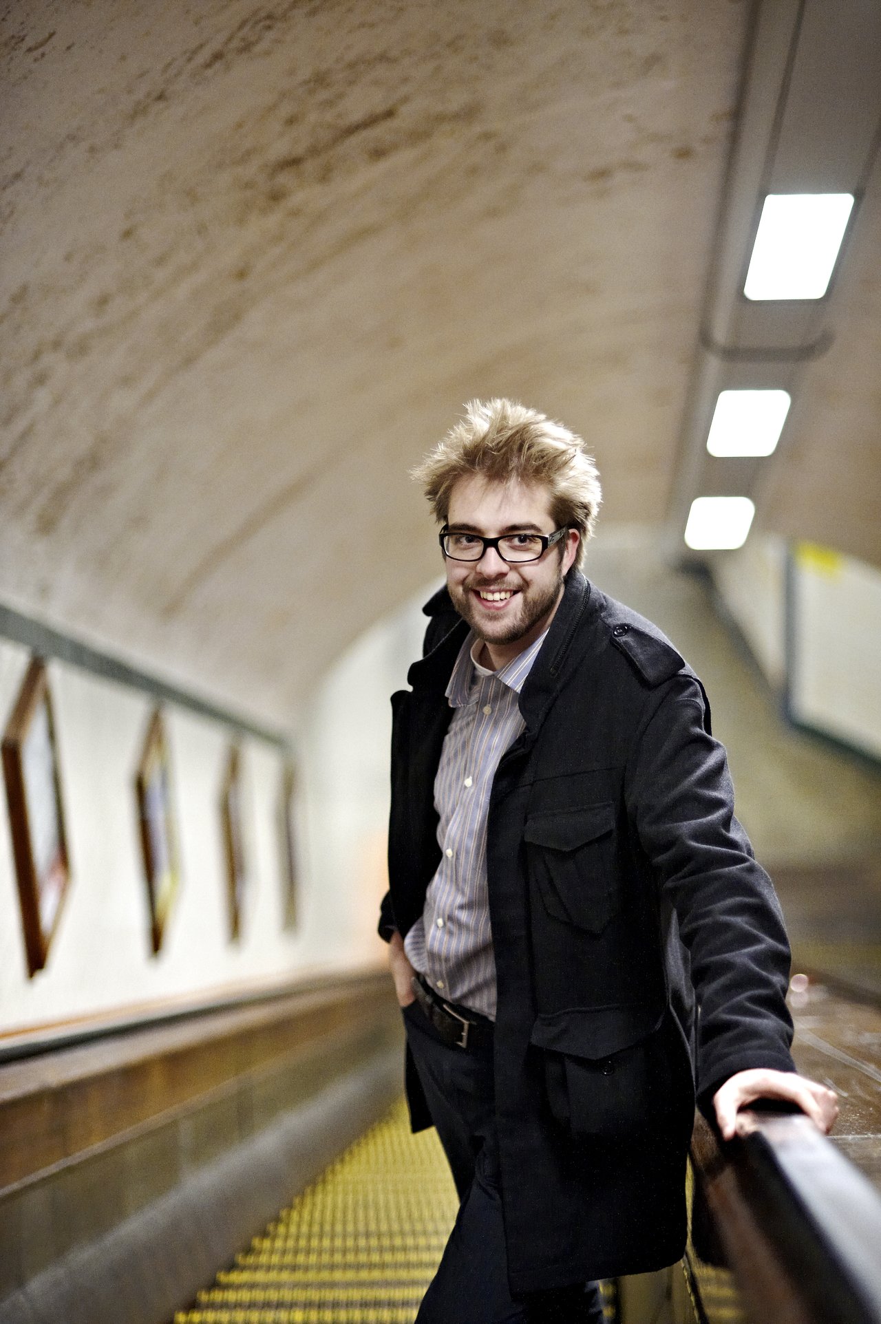 A man with glasses and a black coat smiles while leaning on an escalator handrail in a tunnel.