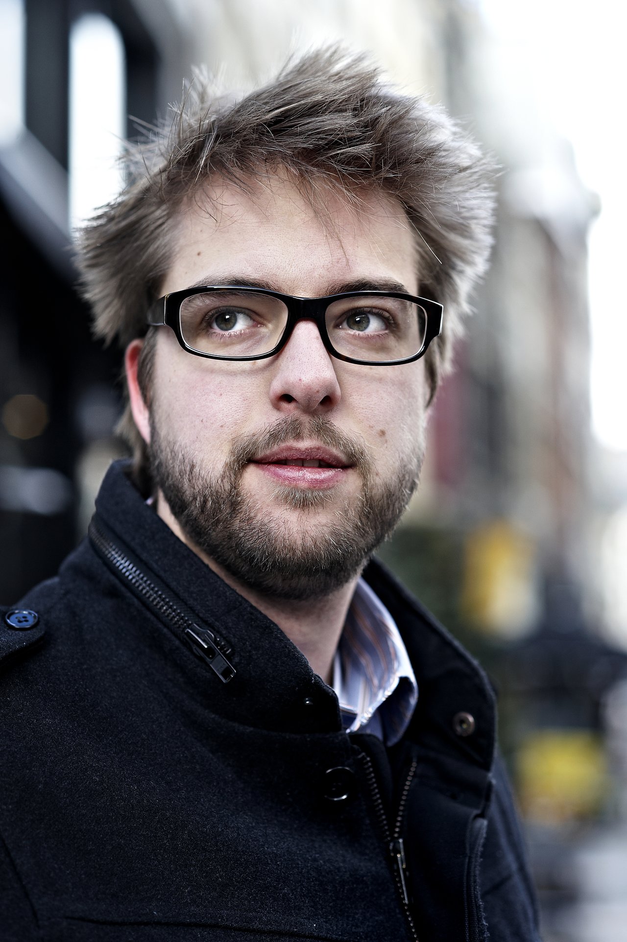 A man with glasses and a beard looks to the side while standing outdoors in a city setting.