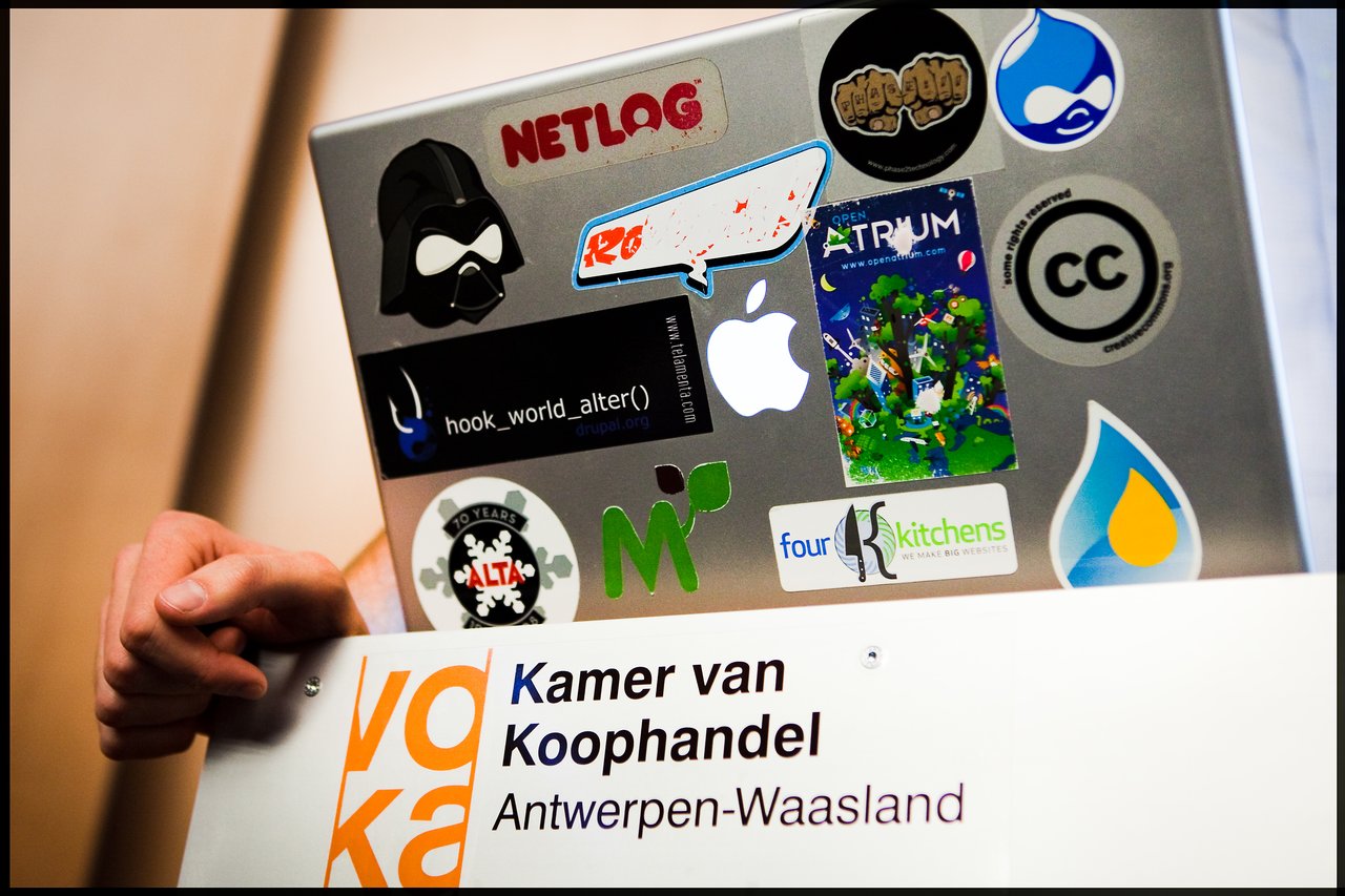 A person holds a laptop covered in stickers behind a Voka Antwerp-Waasland sign during a presentation.
