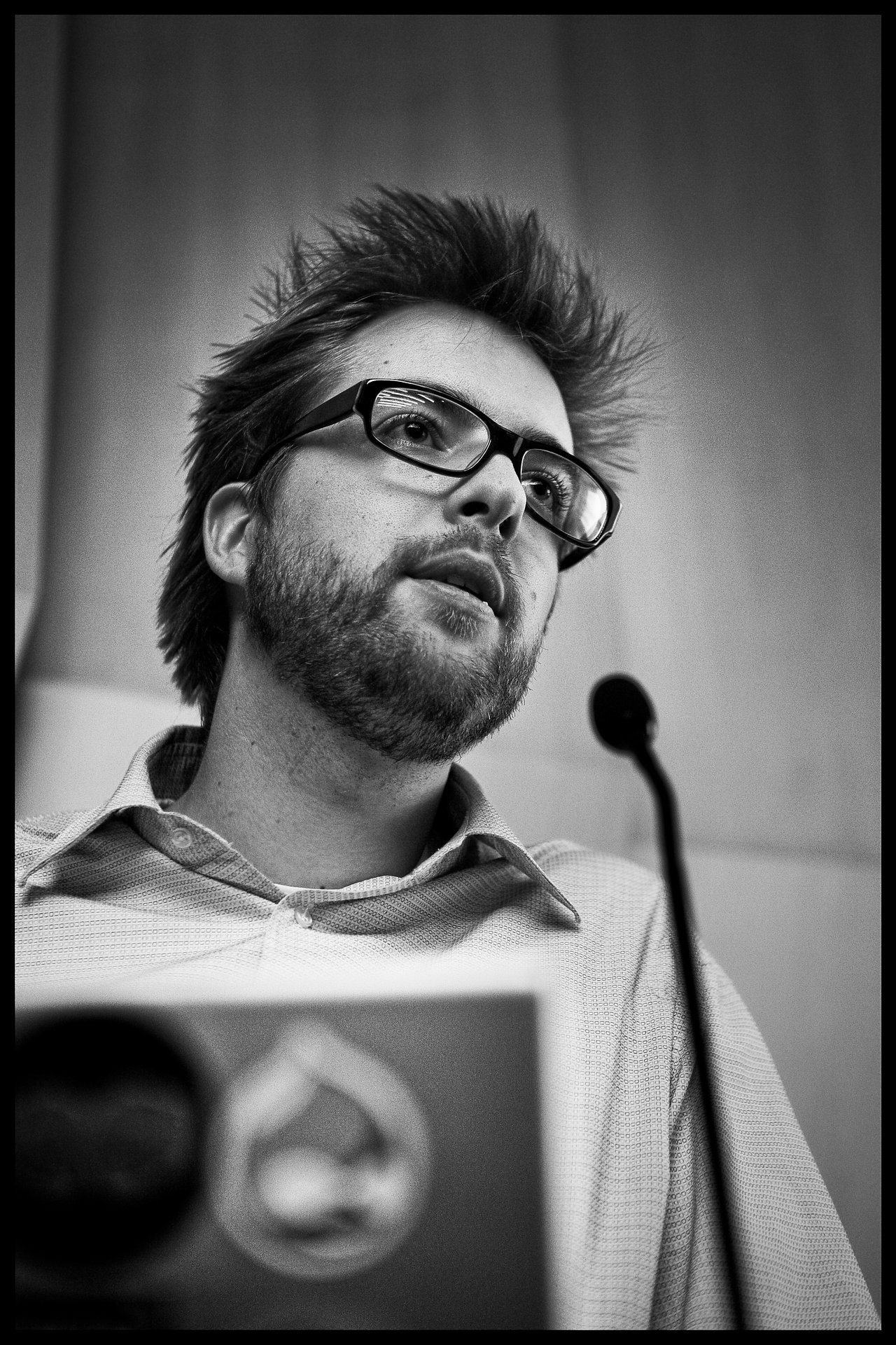 A man with glasses speaks into a microphone during a presentation, with a laptop in front of him.