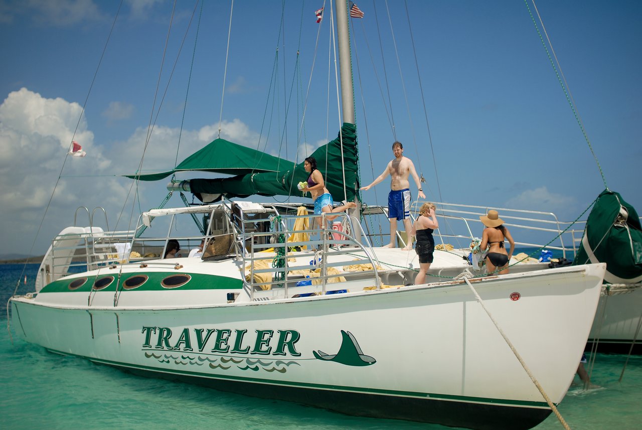 People standing and relaxing on a white catamaran named "Traveler", anchored in clear turquoise water under a sunny sky.