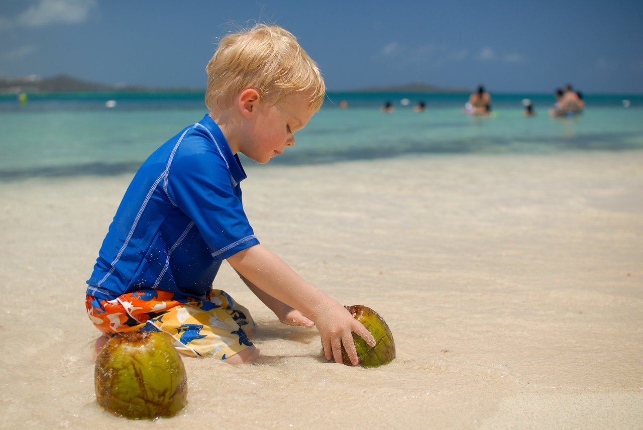 A young child in a blue shirt plays with coconuts in the shallow water at the beach.