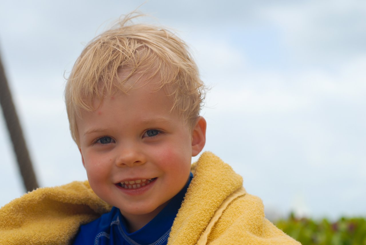 A smiling young child with wet blond hair is wrapped in a yellow towel outdoors.