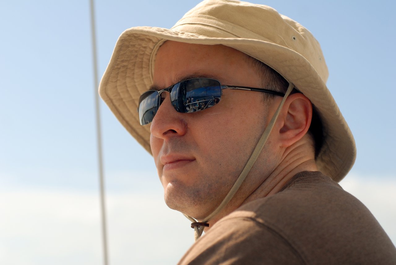 A man wearing sunglasses and a beige sun hat looks off to the side under a bright sky.