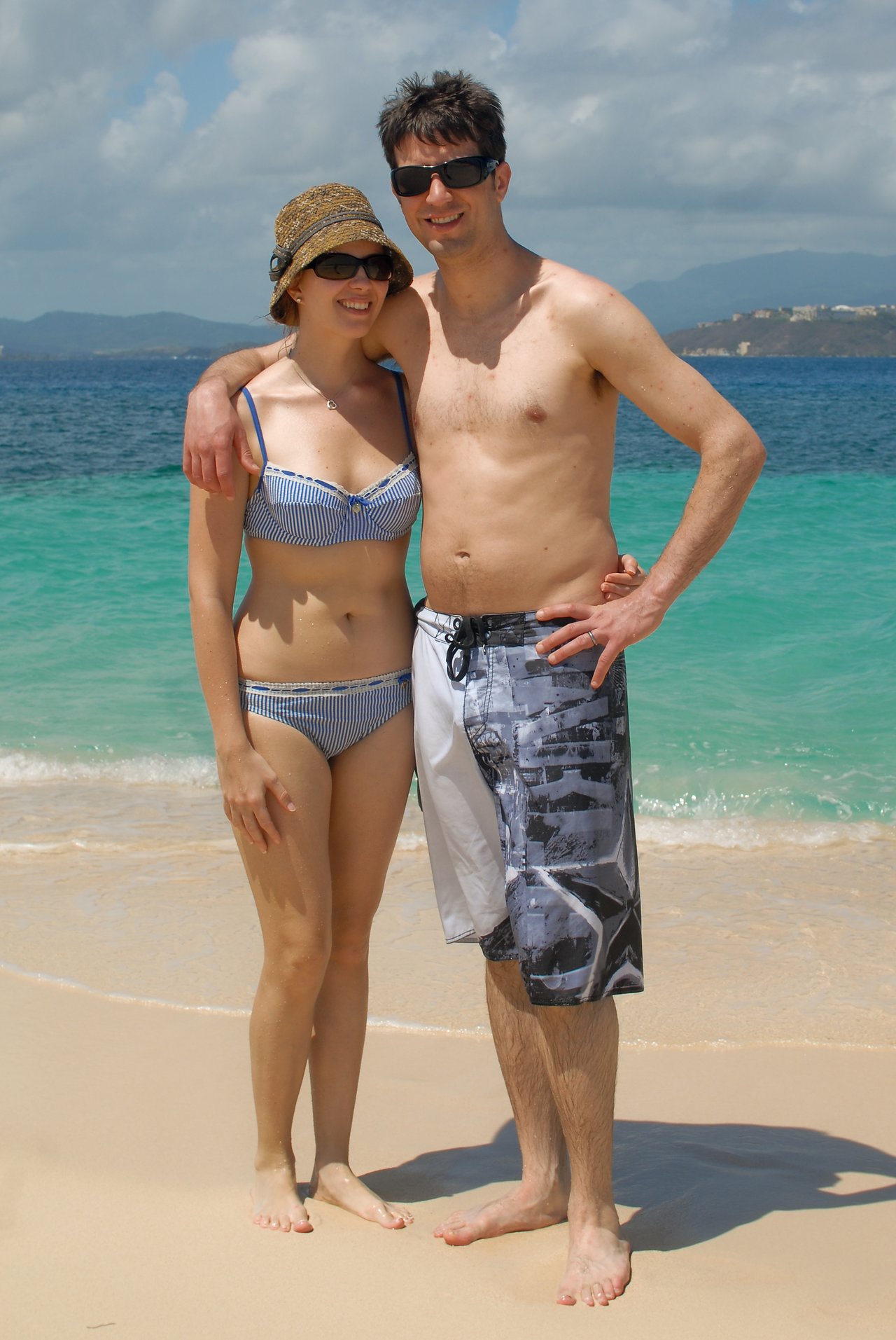 A man and woman in swimsuits stand on a sandy beach, smiling and posing with an arm around each other.