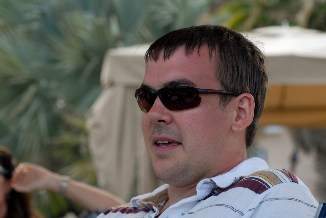 A man wearing sunglasses and a striped polo shirt sits outdoors with a neutral expression.