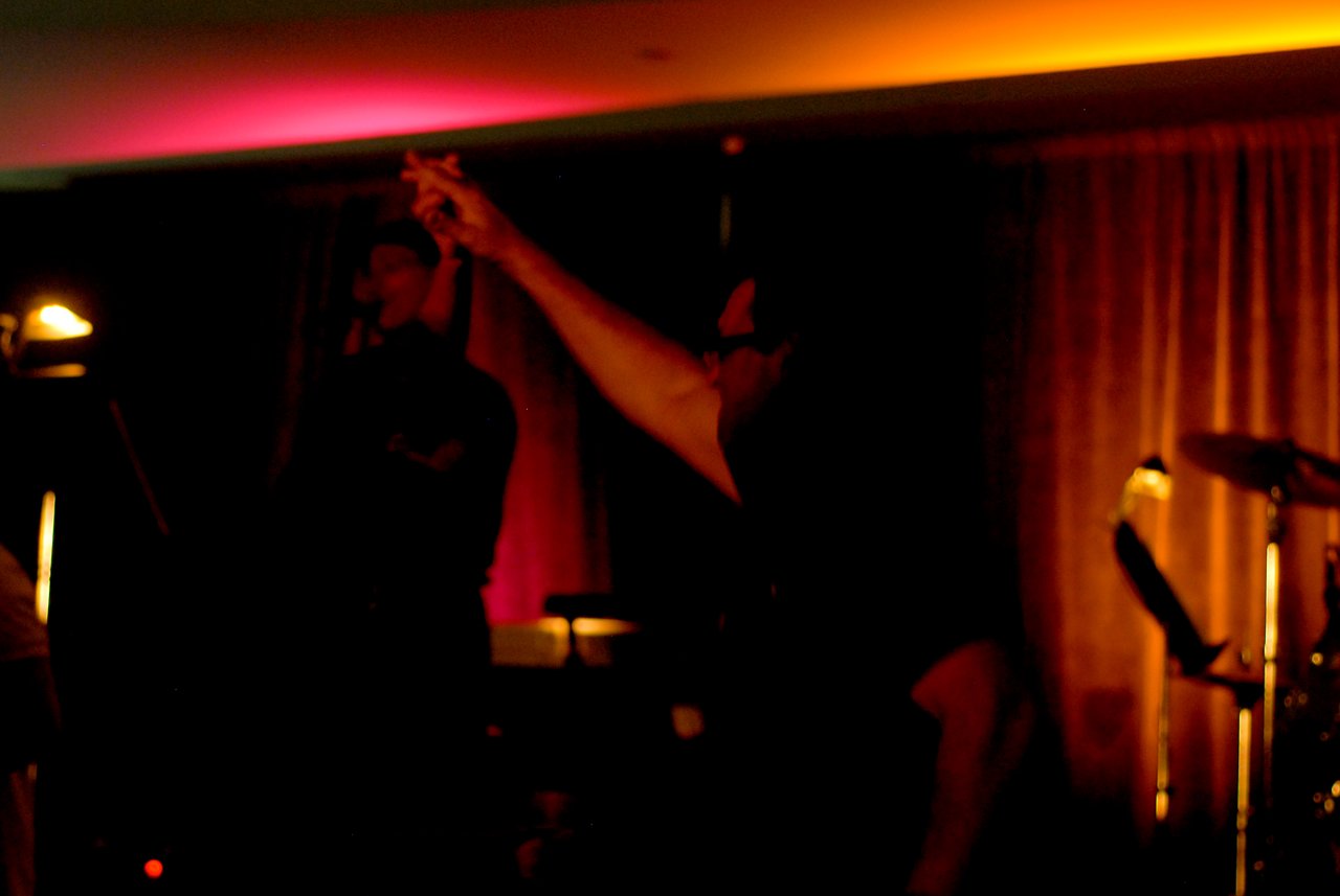 A person dances with their arm raised in a dimly lit room with colorful lighting and a live band.
